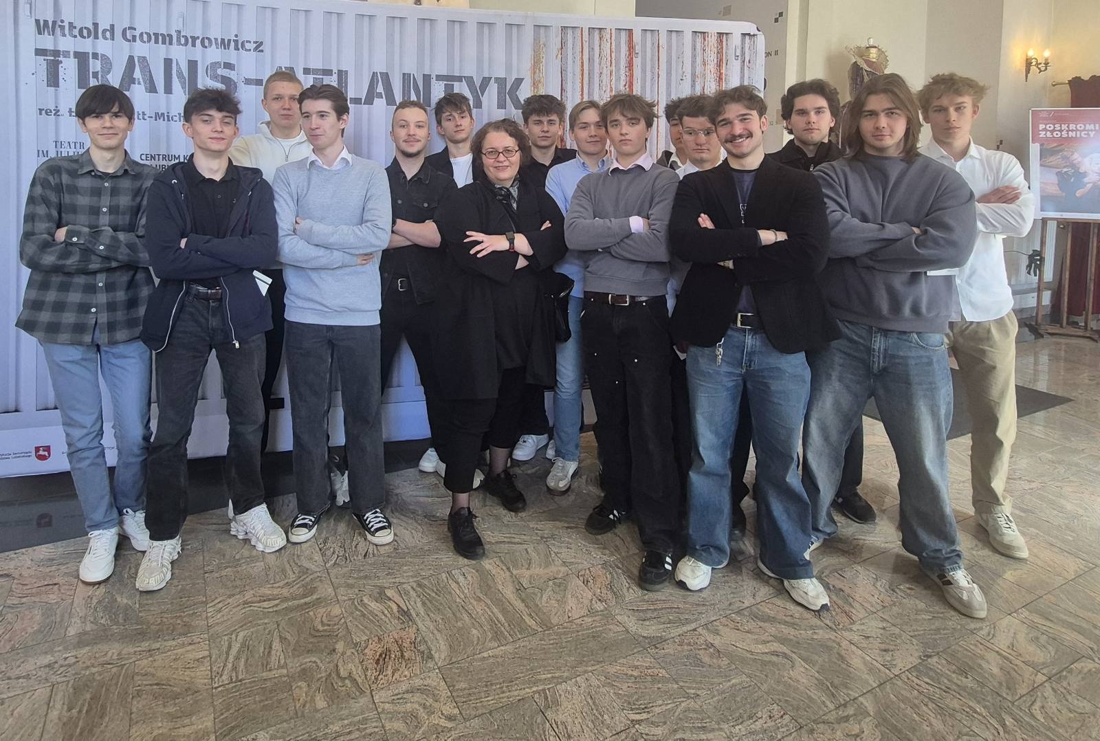 Grupa młodych mężczyzn z nauczycielką pośrodku stoi przed tym samym plakatem; większość ma skrzyżowane ręce i patrzy w stronę aparatu.