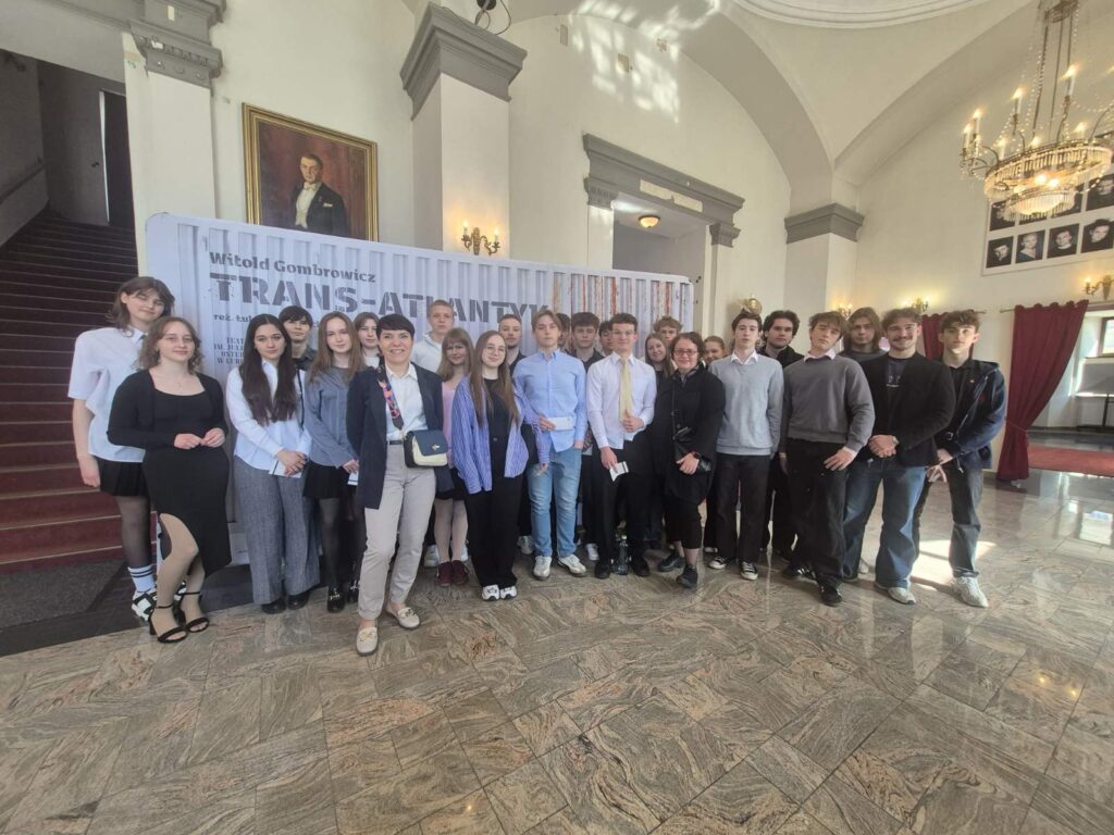 Duża, mieszana grupa uczniów i nauczycieli stoi razem w przestronnym, eleganckim wnętrzu (wysokie sufity, żyrandole), pozując do wspólnego zdjęcia przed plakatem.