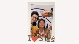 Trzy dorosłe osoby uśmiechają się do zdjęcia zza papierowej ramki z napisem „I ♥ Staś”. Dwie kobiety i mężczyzna pochylają się do środka kadru; jedna z kobiet trzyma pluszowego misia. Wszyscy mają przypinki i koszulki z motywem serca.