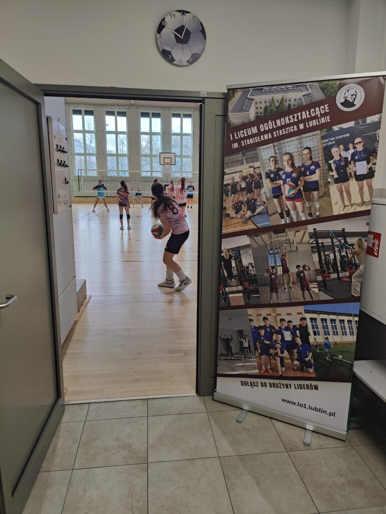 Widok z korytarza prowadzącego do hali sportowej. W otwartych drzwiach widać trwający mecz siatkówki oraz zawodniczkę przygotowującą się do zagrywki. Po prawej stronie stoi wysoki baner promujący I Liceum Ogólnokształcące im. Stanisława Staszica w Lublinie, zawierający zdjęcia uczniów podczas zajęć sportowych i szkolnych wydarzeń. Nad drzwiami znajduje się zegar ścienny z motywem piłki nożnej.