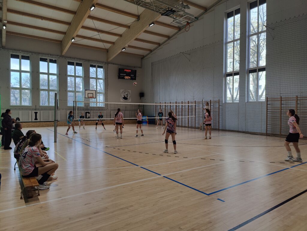 Mecz siatkówki dziewcząt rozgrywany w dużej hali sportowej. Zawodniczki w biało-różowych koszulkach ustawione są po jednej stronie siatki, a drużyna w ciemniejszych strojach po drugiej. Kilka osób siedzi na ławce rezerwowych przy bocznej linii boiska. Hala jest przestronna, z wysokim sufitem, dużymi oknami i elektroniczną tablicą wyników na ścianie.