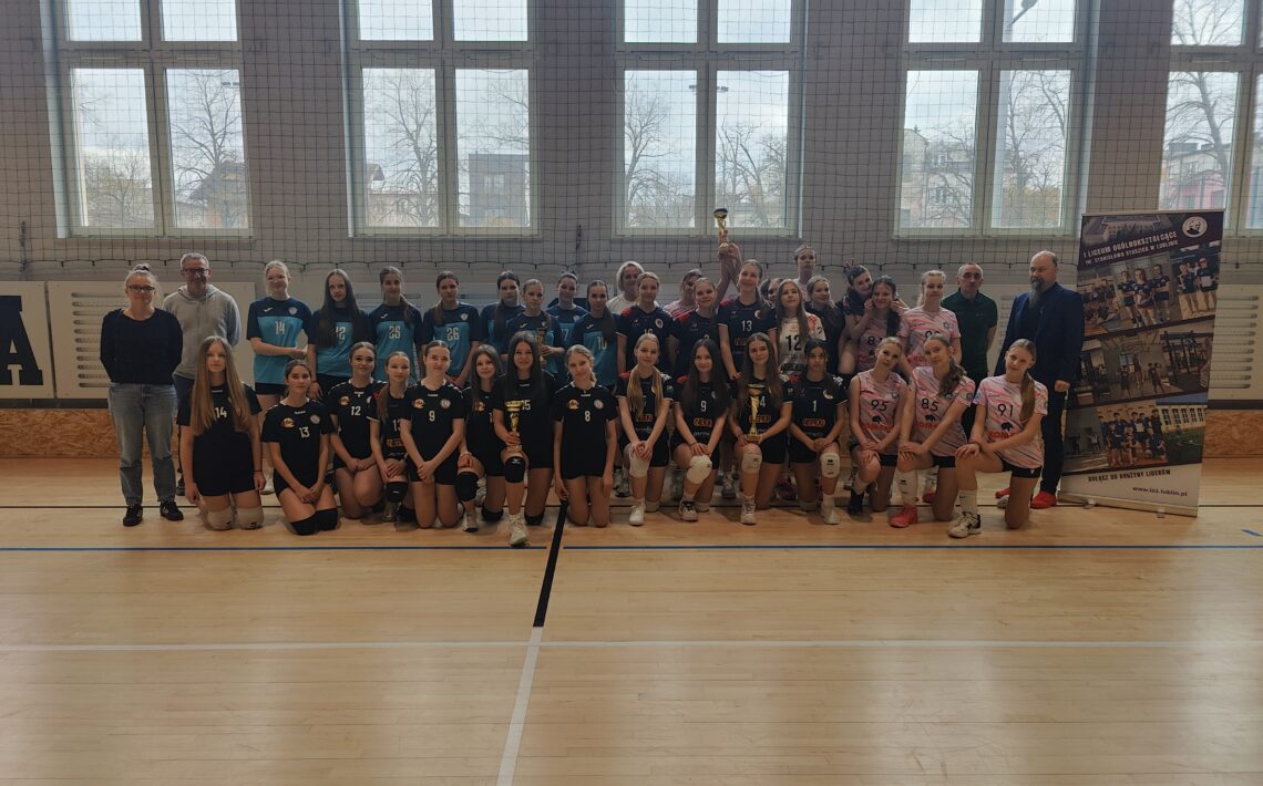 Grupowe zdjęcie drużyn siatkarskich wykonane w hali sportowej. Na środku stoją i klęczą młode zawodniczki w sportowych strojach w kolorach granatowym, turkusowym i biało-różowym. Wśród uczestników znajdują się także trenerzy i opiekunowie. Jedna z zawodniczek trzyma puchar uniesiony do góry. W tle widoczne są wysokie okna hali sportowej oraz baner promujący szkołę.