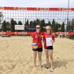 Dwie młode zawodniczki stoją na piaszczystym boisku do siatkówki plażowej przed siatką z czerwonymi taśmami reklamowymi ORLEN. Obie mają czerwone koszulki sportowe i czarne spodenki. Jedna trzyma piłkę siatkową oraz medal zawieszony na szyi, druga prezentuje puchar i dyplom. Dziewczęta pozują do zdjęcia po zakończonych zawodach, a całość ma charakter sportowego sukcesu i wyróżnienia.