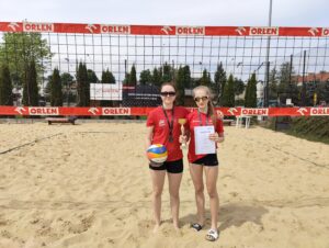 Dwie młode zawodniczki stoją na piaszczystym boisku do siatkówki plażowej przed siatką z czerwonymi taśmami reklamowymi ORLEN. Obie mają czerwone koszulki sportowe i czarne spodenki. Jedna trzyma piłkę siatkową oraz medal zawieszony na szyi, druga prezentuje puchar i dyplom. Dziewczęta pozują do zdjęcia po zakończonych zawodach, a całość ma charakter sportowego sukcesu i wyróżnienia.