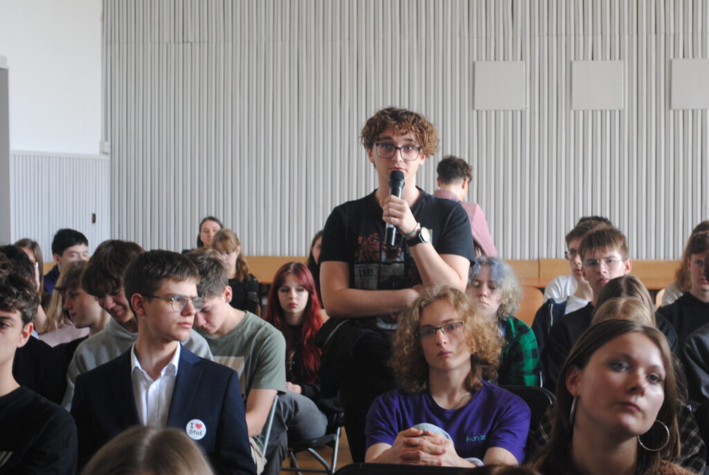 Uczeń przemawia do mikrofonu podczas panelu dyskusyjnego w auli szkolnej.