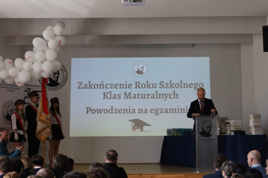 Szerokie ujęcie sali: prowadzący stoi przy podium, przed nim publiczność; po lewej poczet sztandarowy, w tle duży ekran z nazwą uroczystości.
