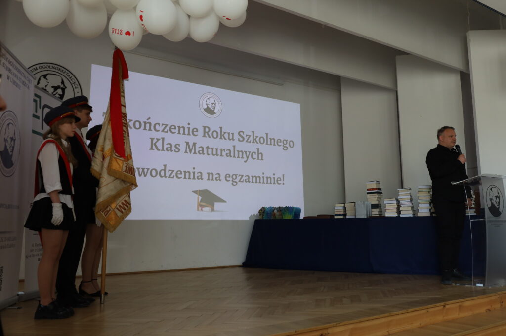 Mężczyzna przemawia przy podium do zgromadzonych; po lewej stoi poczet sztandarowy z flagą, w tle ekran z napisem „Zakończenie Roku Szkolnego Klas Maturalnych. Powodzenia na egzaminie!”.
