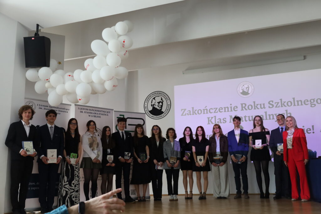 Duża grupa uczniów i nauczycieli stoi na scenie do wspólnego zdjęcia, trzymając książki i nagrody; po prawej widoczna kobieta w czerwonym garniturze.