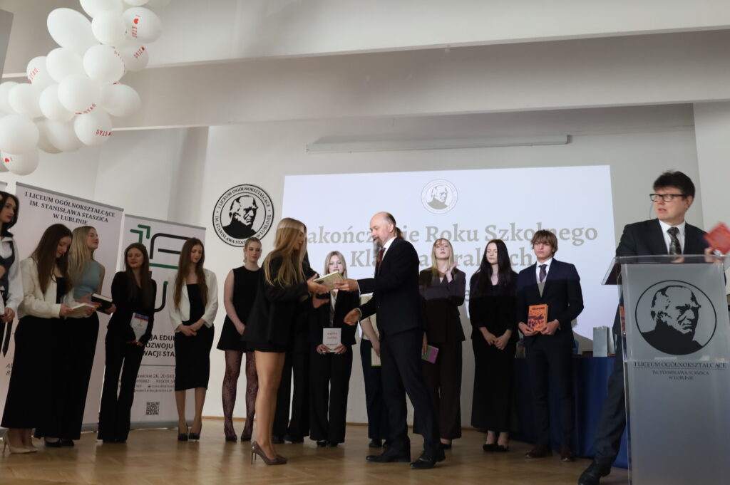 Kolejna uczennica odbiera książkę od nauczyciela; obok stoi prowadzący przy podium oraz grupa uczniów w eleganckich strojach.