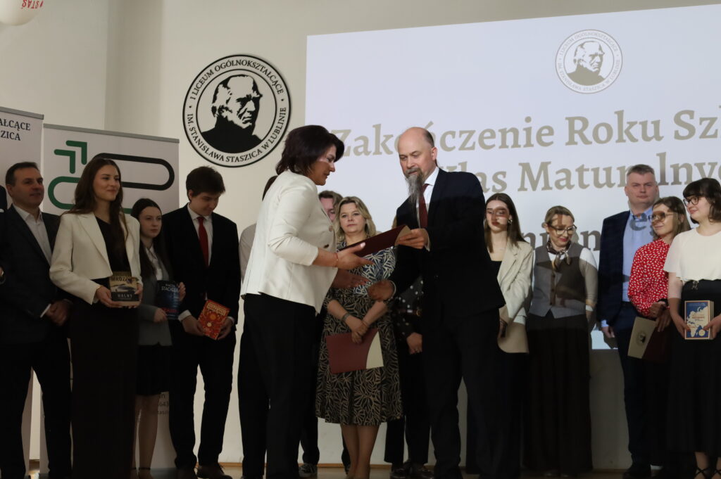 Nauczyciel wręcza dokument kobiecie w jasnej marynarce; wokół stoją uczniowie i nauczyciele, część trzyma książki. W tle ekran z napisem o zakończeniu roku.