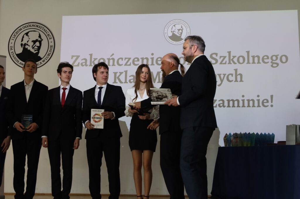 Nauczyciel w ciemnym garniturze wręcza nagrodę ksiązkową