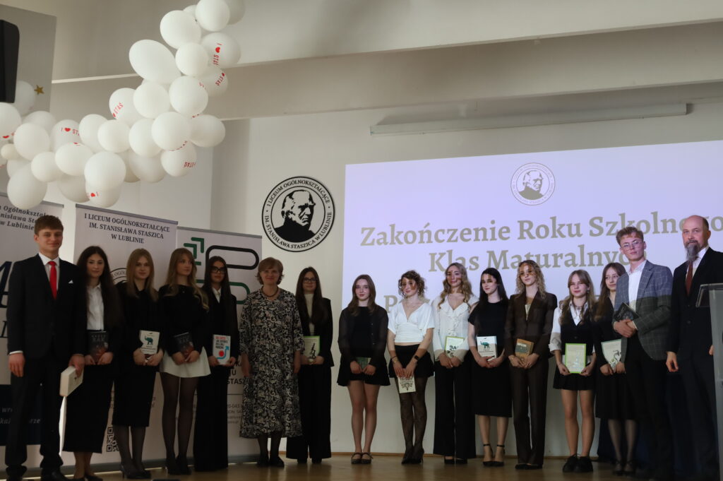 Grupa uczniów, głównie chłopców w garniturach, stoi na scenie z książkami; po prawej nauczyciele obserwują wydarzenie.