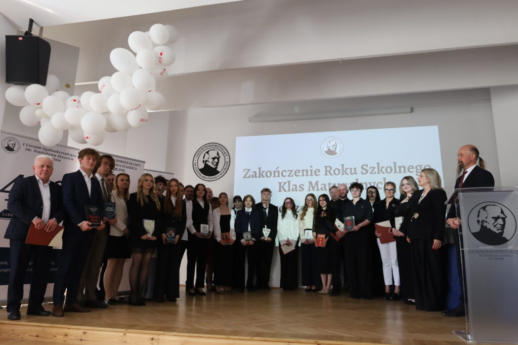 Uczennice i nauczyciele stoją w szeregu, prezentując książki; centralnie widoczna nauczycielka w wzorzystej sukience.