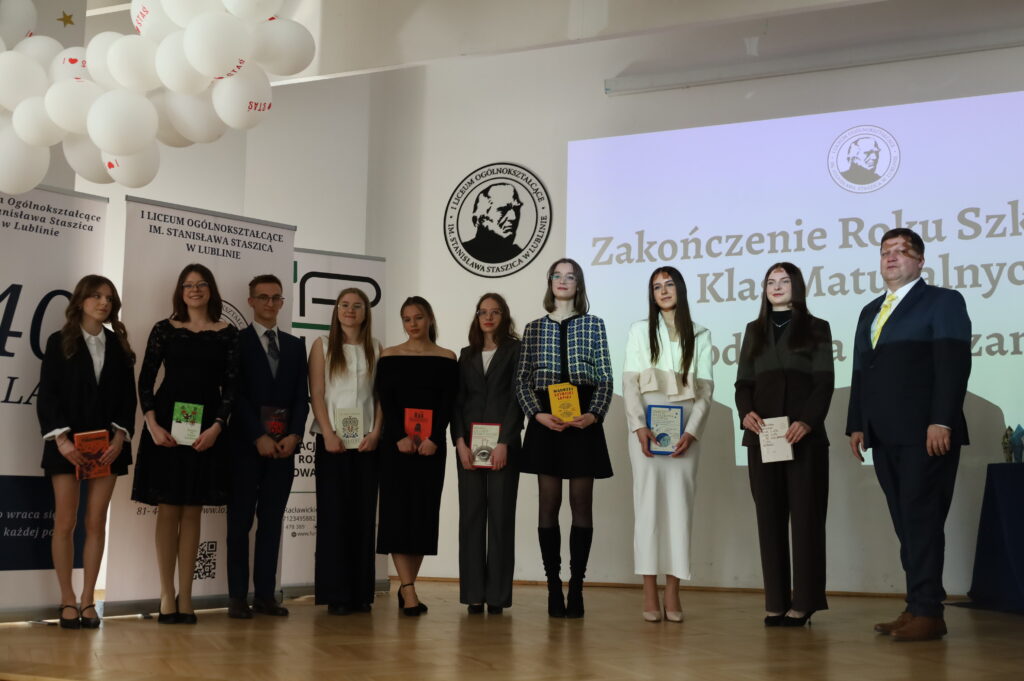 Grupa uczniów i nauczycieli ustawiona do wspólnego zdjęcia na scenie; część trzyma książki i dyplomy, w tle widoczny napis o zakończeniu roku szkolnego.
