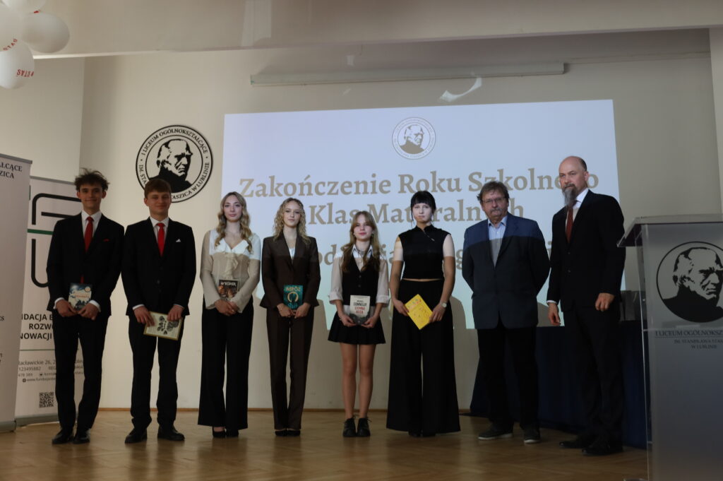 Uczniowie ustawieni w jednej linii pozują do zdjęcia, trzymając książki; obok stoją dwaj nauczyciele. W tle ekran z napisem o zakończeniu roku.