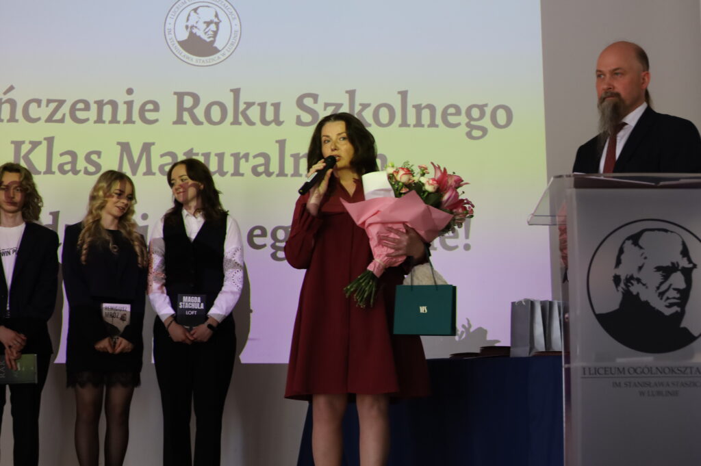 Kobieta w czerwonej sukience przemawia do mikrofonu, trzyma bukiet kwiatów i torbę prezentową. Za nią stoją uczniowie, a po prawej widoczne podium z logo szkoły.