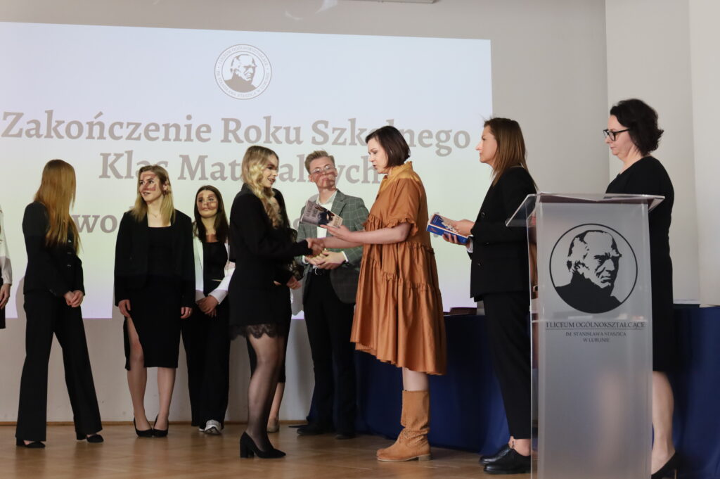 Uczennica odbiera książkę od nauczycielki. Obok stoją inne nauczycielki oraz uczniowie, wszyscy ubrani elegancko.