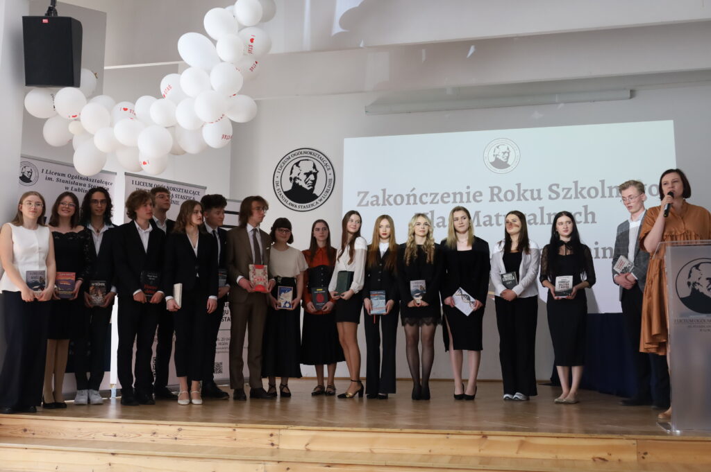 Grupa uczniów stoi na scenie, część trzyma książki (nagrody). Po prawej kobieta przemawia do mikrofonu przy podium.