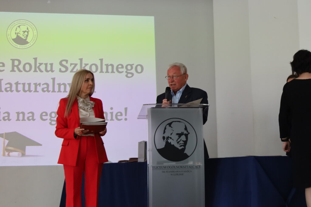 Starszy mężczyzna przemawia przy podium, obok stoi kobieta w czerwonym garniturze trzymająca dokumenty. W tle projekcja z nazwą uroczystości.