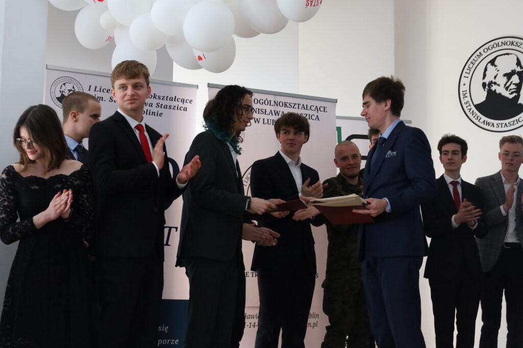 Zbliżenie momentu wręczenia dokumentu uczniowi w garniturze. Wokół inni uczniowie klaszczą, część obserwuje ceremonię.