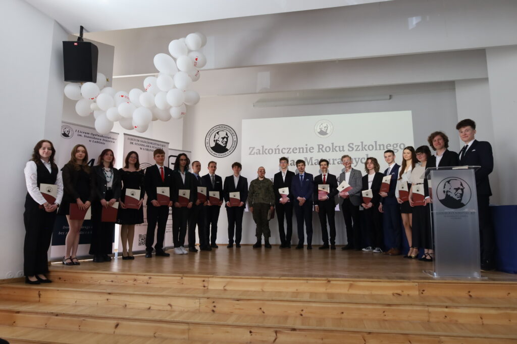 Szerokie ujęcie dużej grupy uczniów na scenie, ustawionych w jednej linii. W tle ekran z napisem o zakończeniu roku i logo szkoły, nad sceną białe balony.