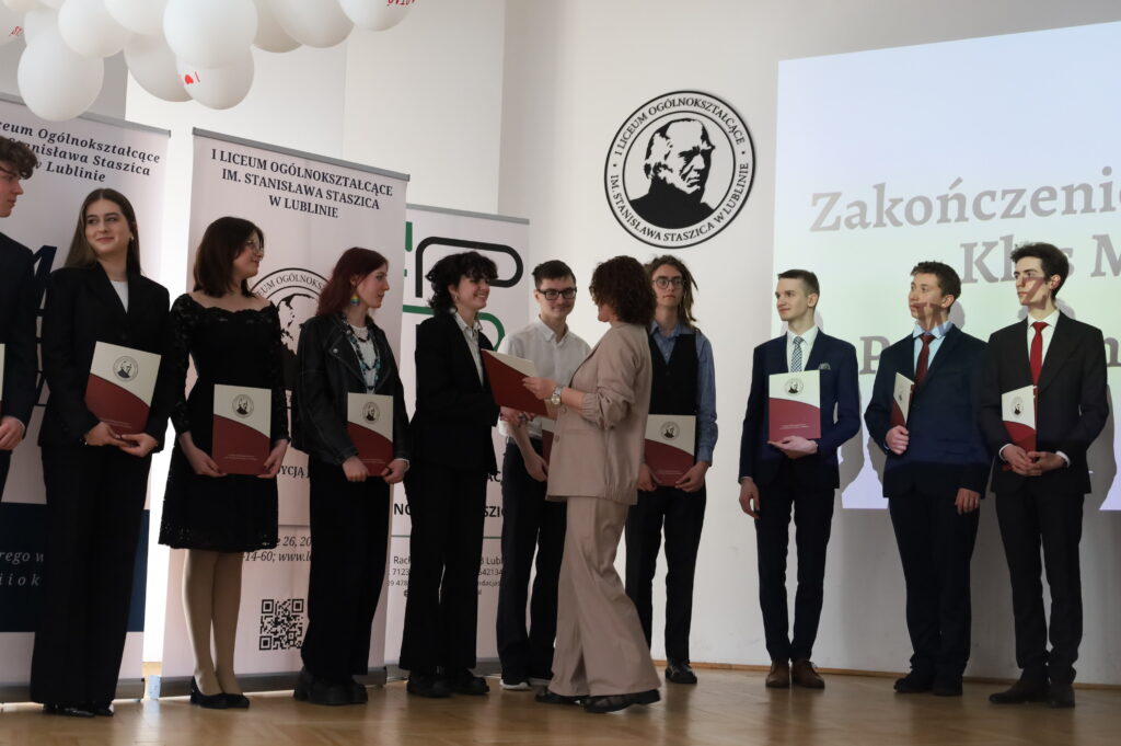 Nauczycielka wręcza dokument uczennicy stojącej w szeregu innych uczniów. Wszyscy ubrani elegancko, część osób trzyma już otrzymane teczki.