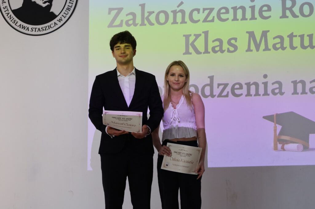 Dwójka uczniów (chłopak w ciemnym garniturze i dziewczyna w białej bluzce) stoi na scenie, trzymając dyplomy. W tle projekcja z napisem „Zakończenie Roku Szkolnego Klas Maturalnych” oraz logo szkoły.