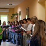 Na korytarzu o jasnych ścianach stoi grupa młodzieży z czarnymi teczkami i śpiewnikami. Uczniowie ustawieni są w półkolu i wyglądają, jakby przygotowywali się do śpiewu lub próby chóru. Po lewej stronie kobieta w zielonej bluzce prowadzi spotkanie lub dyryguje grupą. W tle widoczne są rośliny doniczkowe, półki z książkami i okno wpuszczające naturalne światło. Zdjęcie przedstawia atmosferę wspólnej pracy artystycznej i zaangażowania młodzieży.