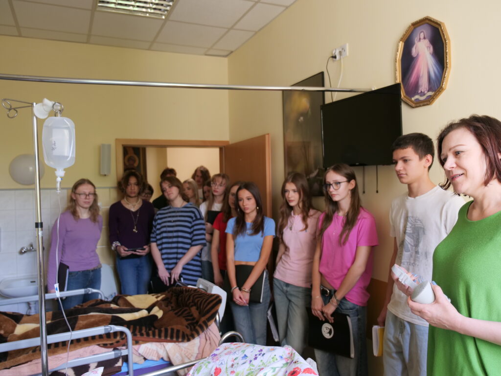 W niewielkim pokoju przypominającym salę opieki lub hospicjum stoi grupa nastolatków słuchających kobiety prowadzącej spotkanie. Po lewej stronie znajduje się łóżko medyczne z kroplówką i kocami. Młodzież ubrana jest swobodnie, część trzyma notesy lub teczki. Na ścianie widoczny jest religijny obraz Jezusa Miłosiernego oraz wyłączony telewizor. Atmosfera zdjęcia jest spokojna i skupiona, sugerująca wizytę edukacyjną lub wolontariacką.