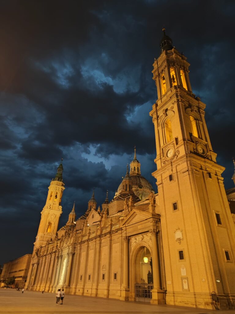 Monumentalna bazylika sfotografowana nocą. Jasno oświetlone wieże i kopuły kontrastują z ciemnym, burzowym niebem. Budowla dominuje nad pustym placem, tworząc imponujący i dramatyczny widok.