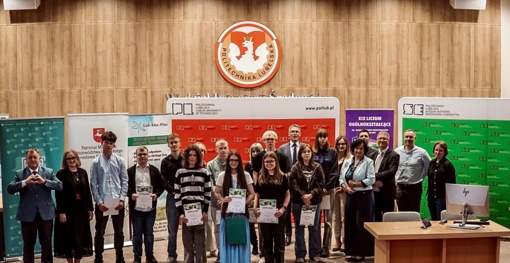 Grupowe zdjęcie wykonane w auli Politechniki Lubelskiej podczas uroczystości związanej z konkursem lub wręczeniem nagród. Na pierwszym planie stoi około dwudziestu osób – głównie młodzież szkolna oraz przedstawiciele organizatorów. Kilku uczestników trzyma dyplomy. W tle widoczne są ścianki promocyjne Politechniki Lubelskiej, partnerów wydarzenia oraz szkoły średniej. Nad grupą znajduje się duże godło Politechniki Lubelskiej umieszczone na drewnianej, akustycznej ścianie. Po prawej stronie stoi biurko z monitorem komputerowym.