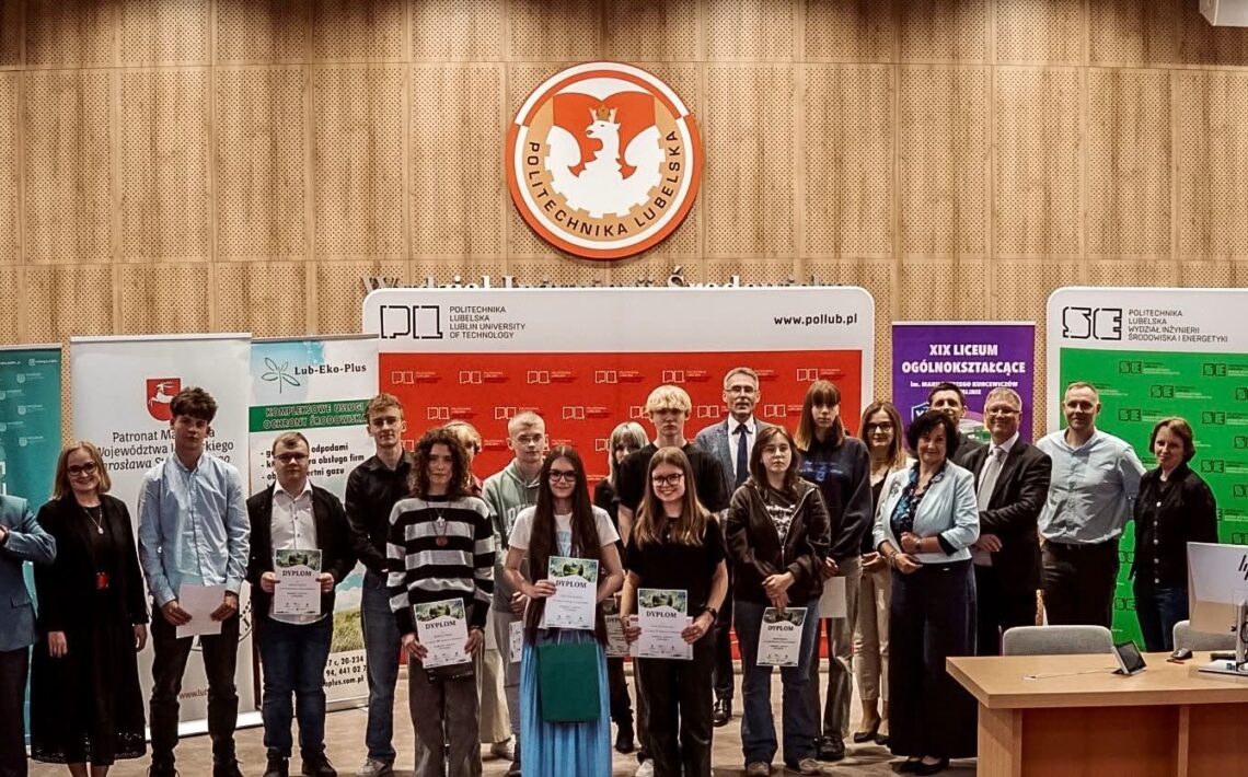 Grupowe zdjęcie wykonane w auli Politechniki Lubelskiej podczas uroczystości związanej z konkursem lub wręczeniem nagród. Na pierwszym planie stoi około dwudziestu osób – głównie młodzież szkolna oraz przedstawiciele organizatorów. Kilku uczestników trzyma dyplomy. W tle widoczne są ścianki promocyjne Politechniki Lubelskiej, partnerów wydarzenia oraz szkoły średniej. Nad grupą znajduje się duże godło Politechniki Lubelskiej umieszczone na drewnianej, akustycznej ścianie. Po prawej stronie stoi biurko z monitorem komputerowym.