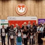 Grupowe zdjęcie wykonane w auli Politechniki Lubelskiej podczas uroczystości związanej z konkursem lub wręczeniem nagród. Na pierwszym planie stoi około dwudziestu osób – głównie młodzież szkolna oraz przedstawiciele organizatorów. Kilku uczestników trzyma dyplomy. W tle widoczne są ścianki promocyjne Politechniki Lubelskiej, partnerów wydarzenia oraz szkoły średniej. Nad grupą znajduje się duże godło Politechniki Lubelskiej umieszczone na drewnianej, akustycznej ścianie. Po prawej stronie stoi biurko z monitorem komputerowym.