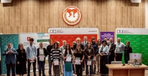 Grupowe zdjęcie wykonane w auli Politechniki Lubelskiej podczas uroczystości związanej z konkursem lub wręczeniem nagród. Na pierwszym planie stoi około dwudziestu osób – głównie młodzież szkolna oraz przedstawiciele organizatorów. Kilku uczestników trzyma dyplomy. W tle widoczne są ścianki promocyjne Politechniki Lubelskiej, partnerów wydarzenia oraz szkoły średniej. Nad grupą znajduje się duże godło Politechniki Lubelskiej umieszczone na drewnianej, akustycznej ścianie. Po prawej stronie stoi biurko z monitorem komputerowym.
