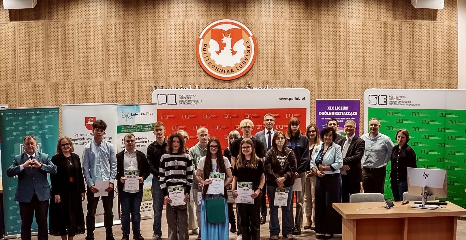 Grupowe zdjęcie wykonane w auli Politechniki Lubelskiej podczas uroczystości związanej z konkursem lub wręczeniem nagród. Na pierwszym planie stoi około dwudziestu osób – głównie młodzież szkolna oraz przedstawiciele organizatorów. Kilku uczestników trzyma dyplomy. W tle widoczne są ścianki promocyjne Politechniki Lubelskiej, partnerów wydarzenia oraz szkoły średniej. Nad grupą znajduje się duże godło Politechniki Lubelskiej umieszczone na drewnianej, akustycznej ścianie. Po prawej stronie stoi biurko z monitorem komputerowym.