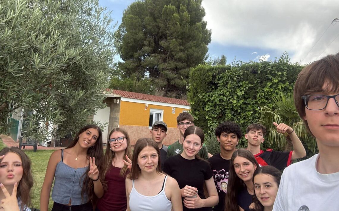 Pamiątkowe selfie grupy młodzieży wykonane w ogrodzie. Kilkanaście osób uśmiecha się do aparatu, niektórzy pokazują gest zwycięstwa lub pozują w zabawny sposób. W tle znajdują się drzewa, żywopłot oraz budynek mieszkalny.