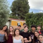 Pamiątkowe selfie grupy młodzieży wykonane w ogrodzie. Kilkanaście osób uśmiecha się do aparatu, niektórzy pokazują gest zwycięstwa lub pozują w zabawny sposób. W tle znajdują się drzewa, żywopłot oraz budynek mieszkalny.