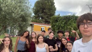 Pamiątkowe selfie grupy młodzieży wykonane w ogrodzie. Kilkanaście osób uśmiecha się do aparatu, niektórzy pokazują gest zwycięstwa lub pozują w zabawny sposób. W tle znajdują się drzewa, żywopłot oraz budynek mieszkalny.