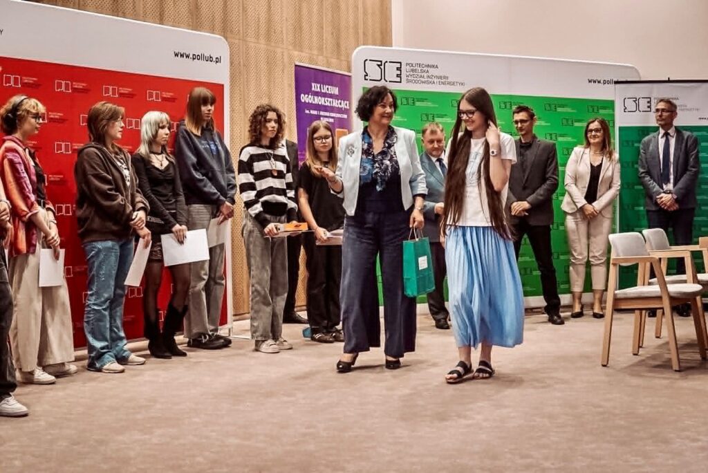Moment wręczania nagrody podczas uroczystości na Politechnice Lubelskiej. Na środku kadru młoda uczestniczka w białej koszulce i długiej, jasnoniebieskiej spódnicy odbiera upominek od kobiety w jasnym żakiecie. W tle stoi grupa pozostałych uczestników oraz organizatorów wydarzenia. Za nimi znajdują się kolorowe ścianki promocyjne Politechniki Lubelskiej i szkoły średniej. Atmosfera zdjęcia jest oficjalna, ale serdeczna i uroczysta.
