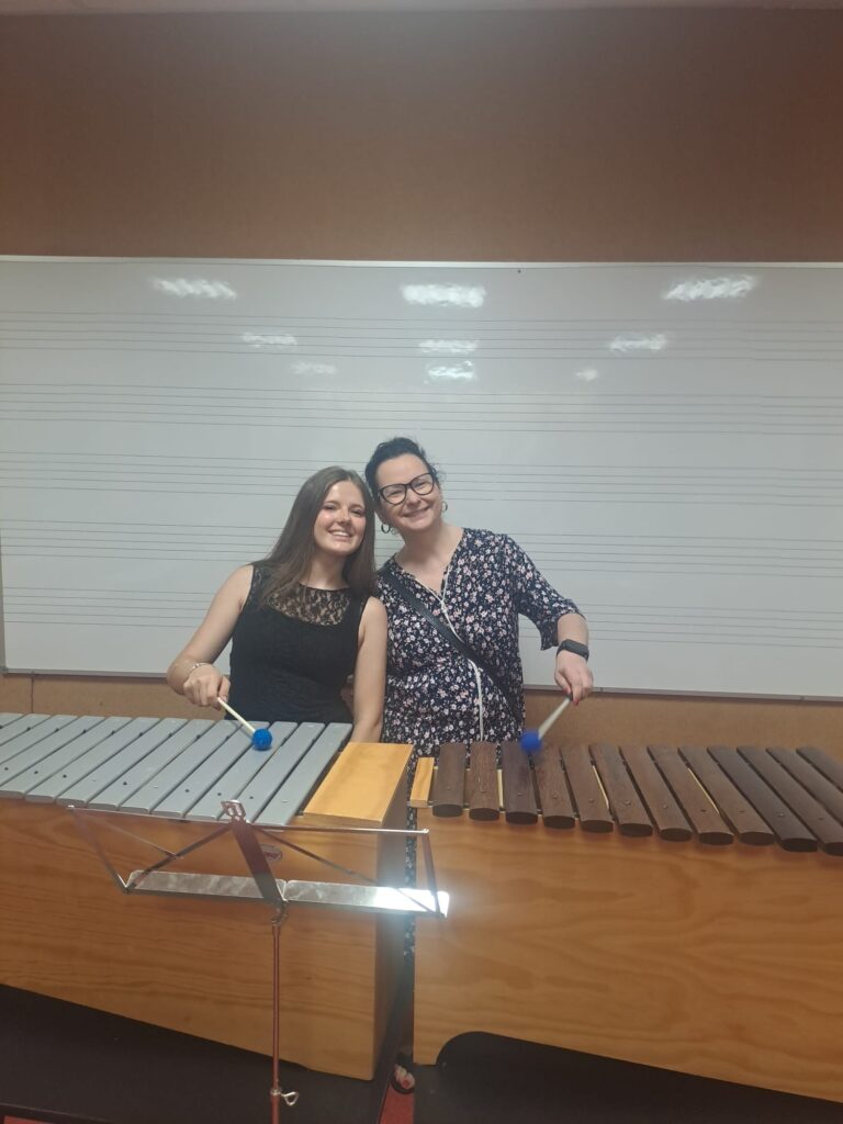 Dwie osoby stoją przy instrumentach perkusyjnych i trzymają pałeczki do gry. Uśmiechają się do zdjęcia na tle sali muzycznej z dużą tablicą do zapisu nut. Kadr pokazuje wspólną aktywność muzyczną.