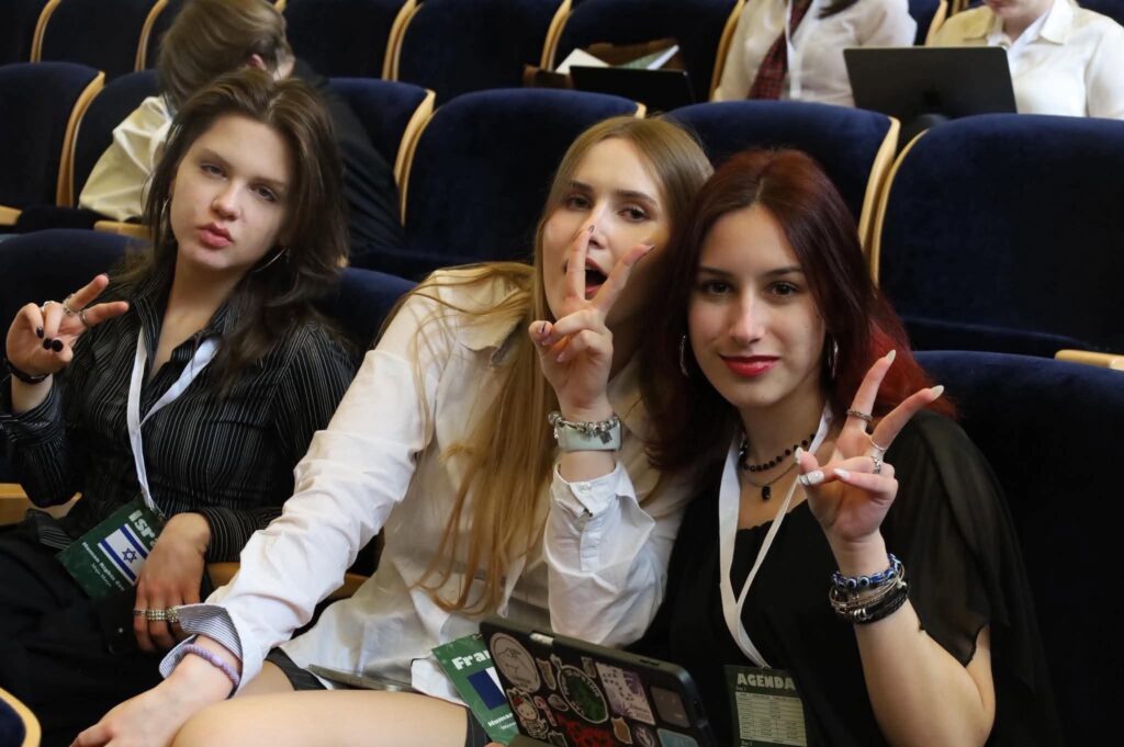 Trzy uczestniczki siedzą na widowni konferencyjnej i pozują do zdjęcia. Uśmiechają się do aparatu oraz pokazują gest „V”, mając na szyjach identyfikatory uczestniczek wydarzenia.