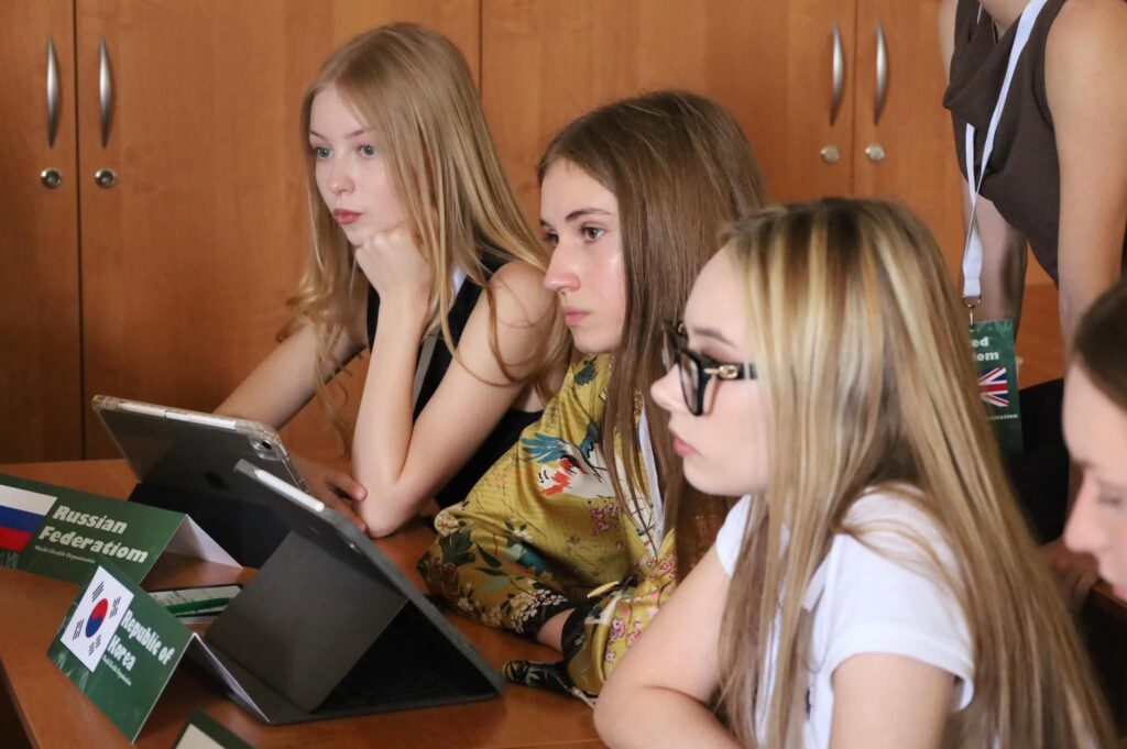 Uczestniczki debaty pracują przy stole konferencyjnym z tabletami i materiałami roboczymi. Przed nimi znajdują się tabliczki reprezentowanych państw, a atmosfera wskazuje na trwające obrady i analizę omawianych tematów.