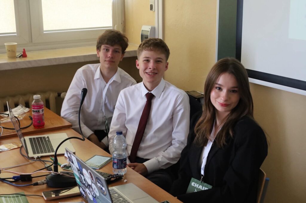 Trzech uczestników pełniących funkcje organizacyjne lub przewodniczących obrad siedzi przy stanowisku wyposażonym w laptopy i sprzęt konferencyjny. Pozują do zdjęcia podczas pracy nad przebiegiem sesji.