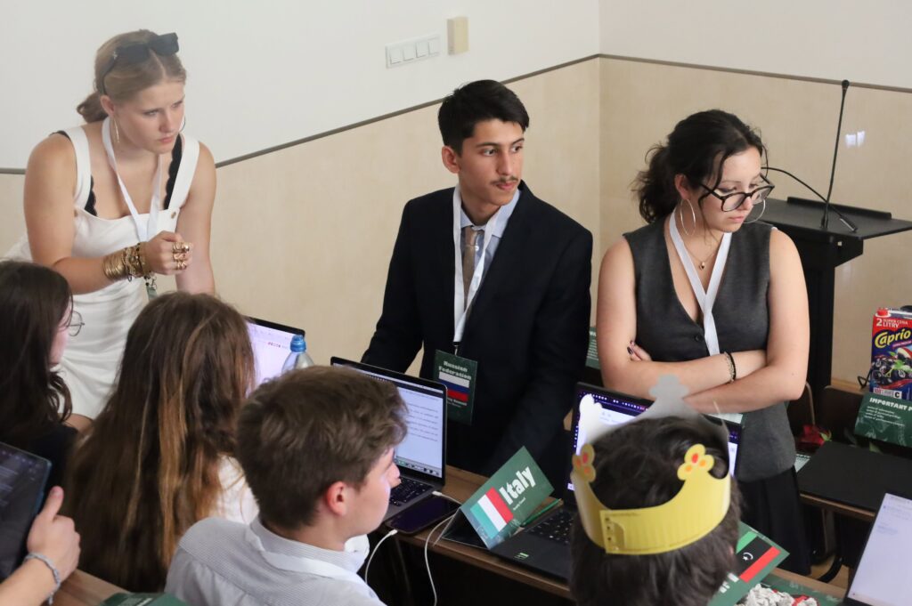 Uczestnicy międzynarodowej symulacji debat prowadzą dyskusję przy stołach konferencyjnych. Widoczne są laptopy, identyfikatory oraz tabliczki państw, co podkreśla formalny charakter spotkania.