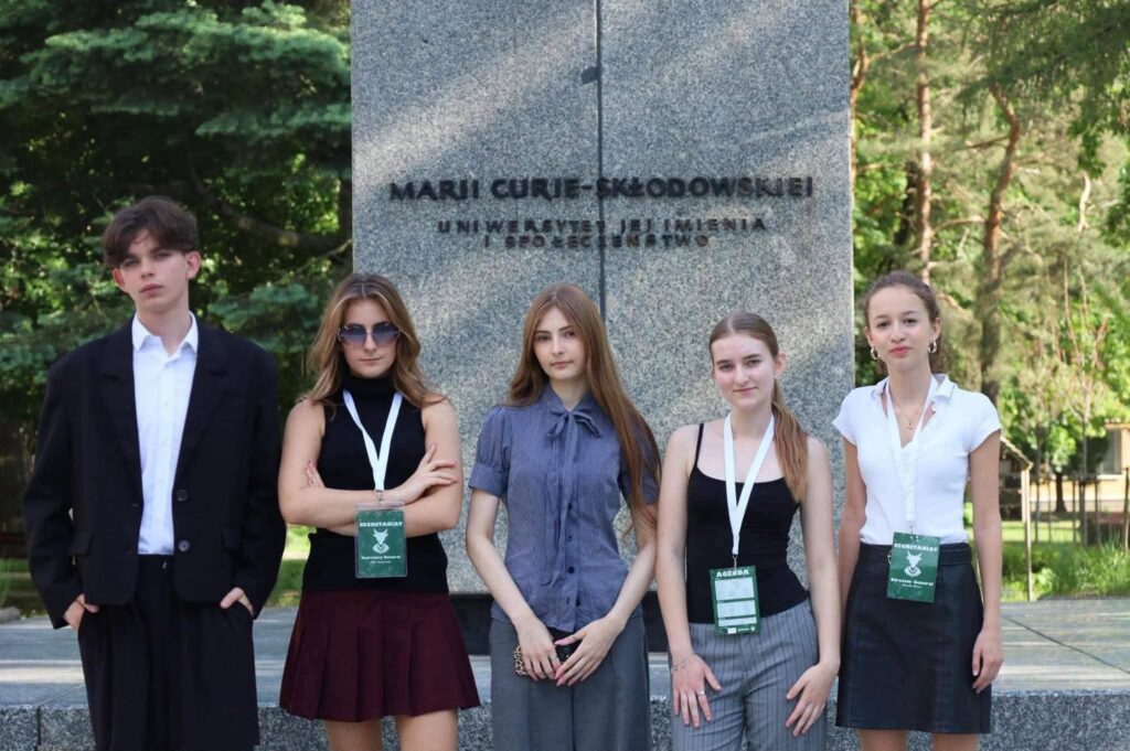 Pięcioro uczestników wydarzenia pozuje do pamiątkowego zdjęcia przed pomnikiem Marii Curie-Skłodowskiej. Wszyscy mają identyfikatory konferencyjne, a fotografia została wykonana na terenie kampusu w otoczeniu zieleni.