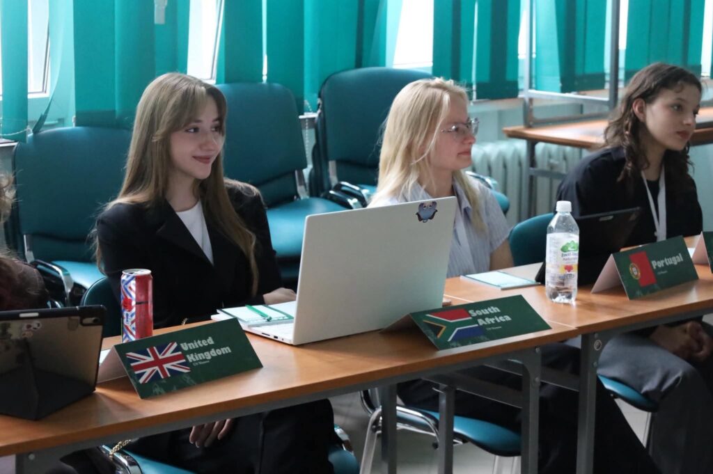 Uczestniczki reprezentujące różne państwa siedzą przy stole konferencyjnym z laptopami i notatkami. Przed nimi ustawiono tabliczki z nazwami krajów, a zdjęcie przedstawia moment obrad.