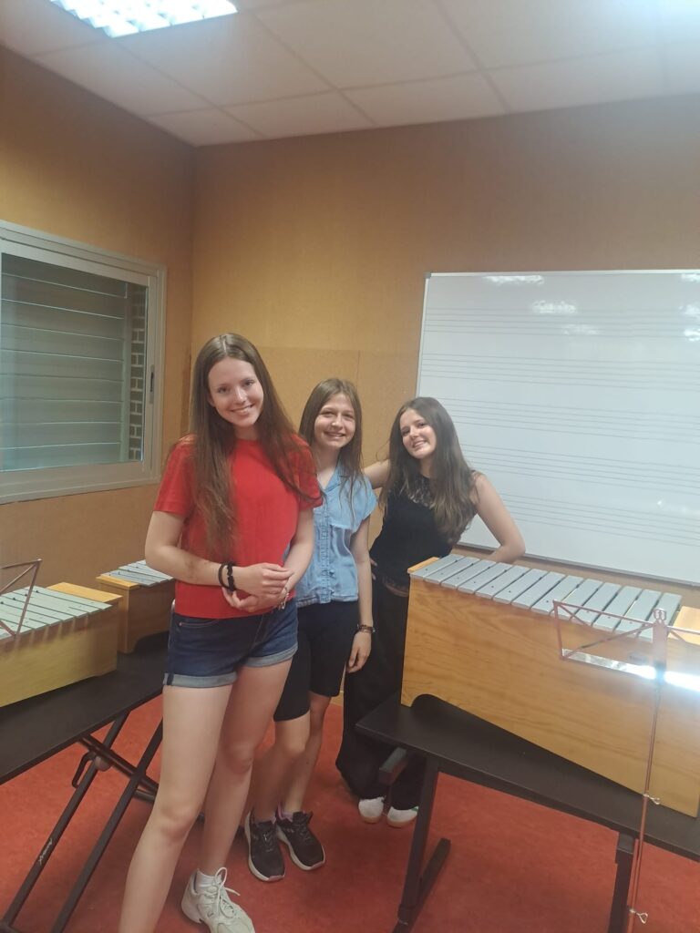Trzy nastolatki stoją w sali muzycznej pomiędzy instrumentami perkusyjnymi przypominającymi ksylofony lub marimby. Uśmiechają się do aparatu, a w tle widoczna jest tablica z pięciolinią. Zdjęcie dokumentuje zajęcia lub warsztaty muzyczne.