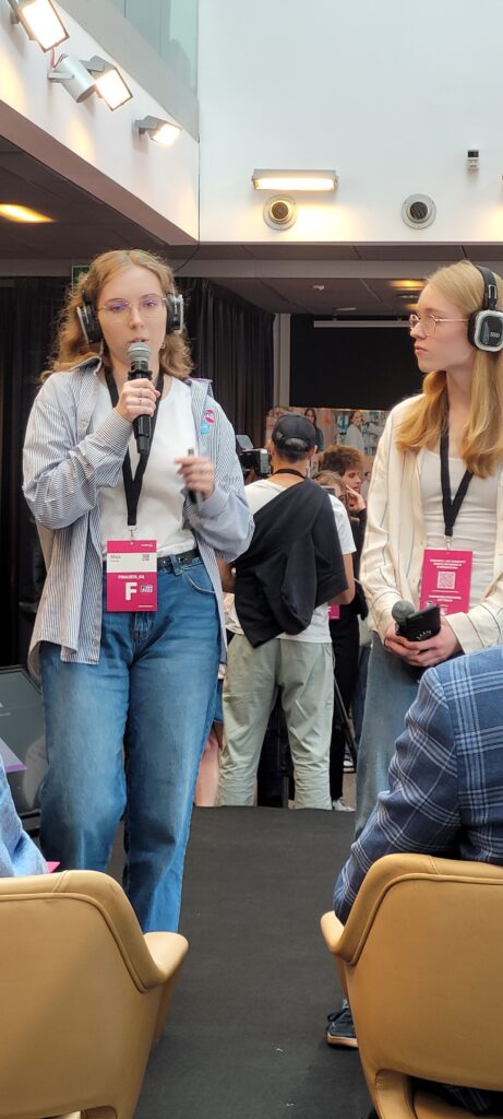 Dwie finalistki podczas wystąpienia w sali konferencyjnej. Jedna mówi do mikrofonu, druga słucha, trzymając mikrofon w dłoni. Obie mają słuchawki i identyfikatory uczestniczek. W tle widoczna jest publiczność.