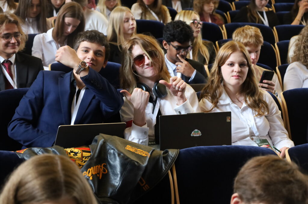 Uczestnicy siedzą na widowni konferencyjnej podczas sesji. Część z nich pracuje na laptopach, inni obserwują wydarzenie, a kilka osób pozuje do zdjęcia w swobodnej atmosferze.