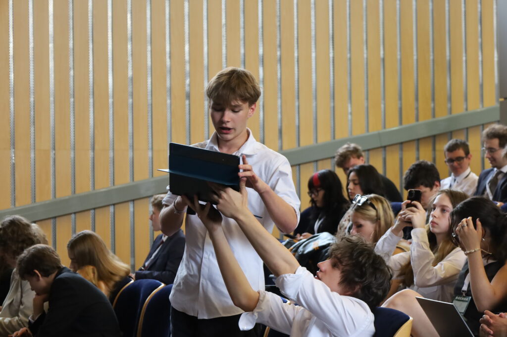 Dwóch uczestników współpracuje przy obsłudze tabletu podczas obrad na sali konferencyjnej. Wokół nich siedzą inni uczestnicy wyposażeni w laptopy i telefony.